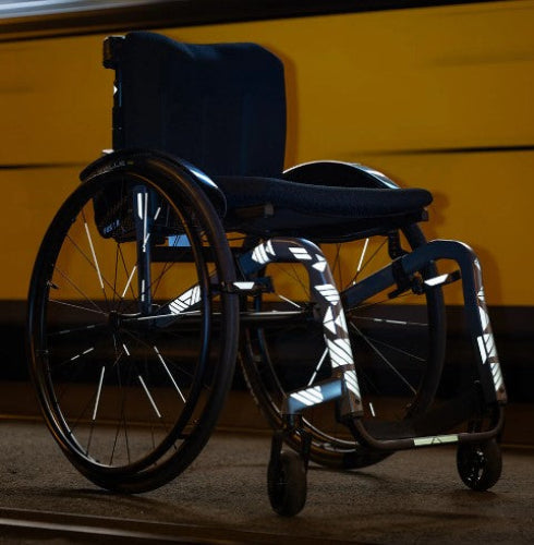 A wheelchair with reflective tape on its frame is positioned on a surface, with a yellow background behind it.