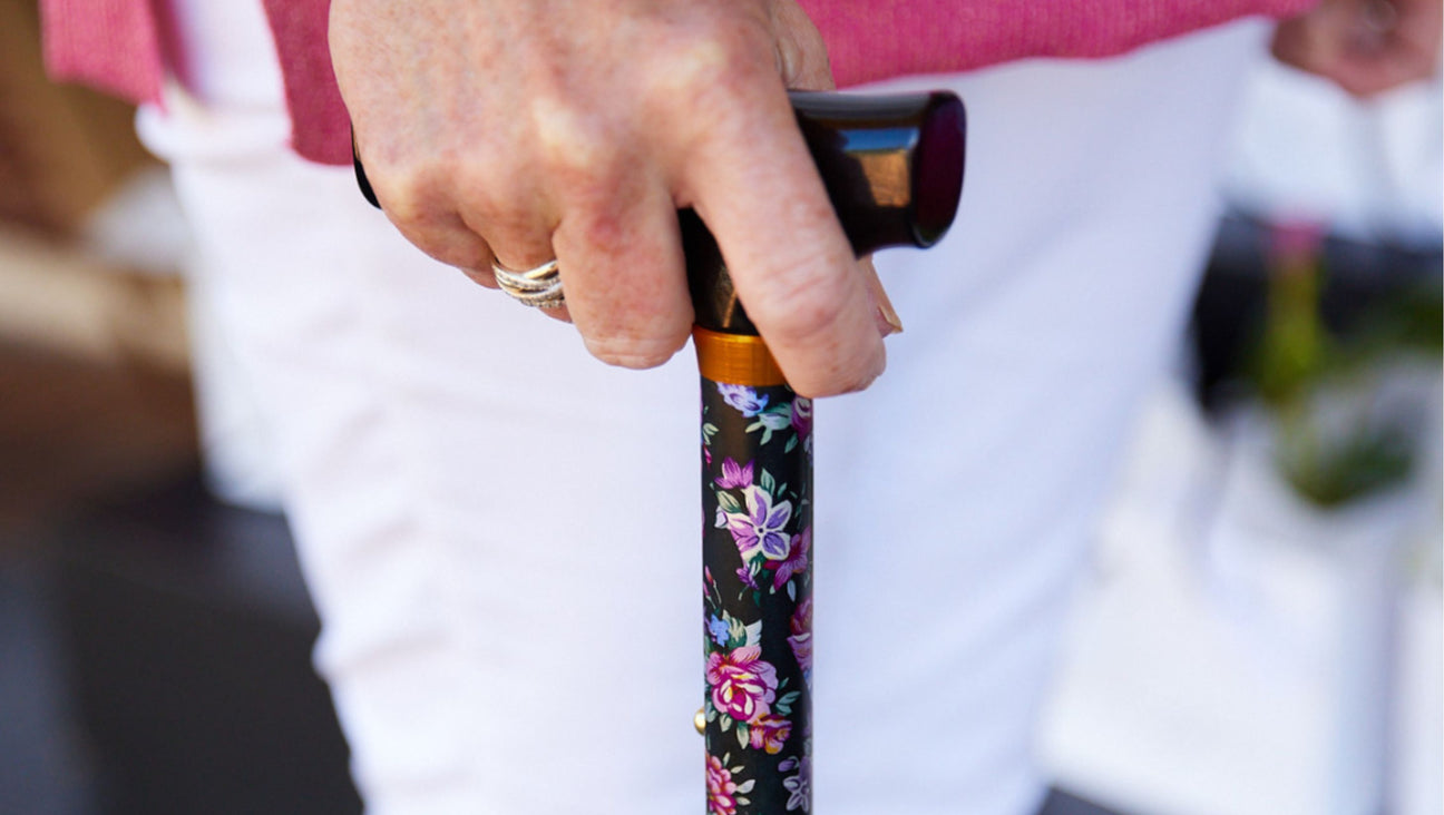 A close-up of a hand holding a black cane decorated with colorful floral patterns. The person is wearing white pants, a pink top, and multiple rings on their fingers.