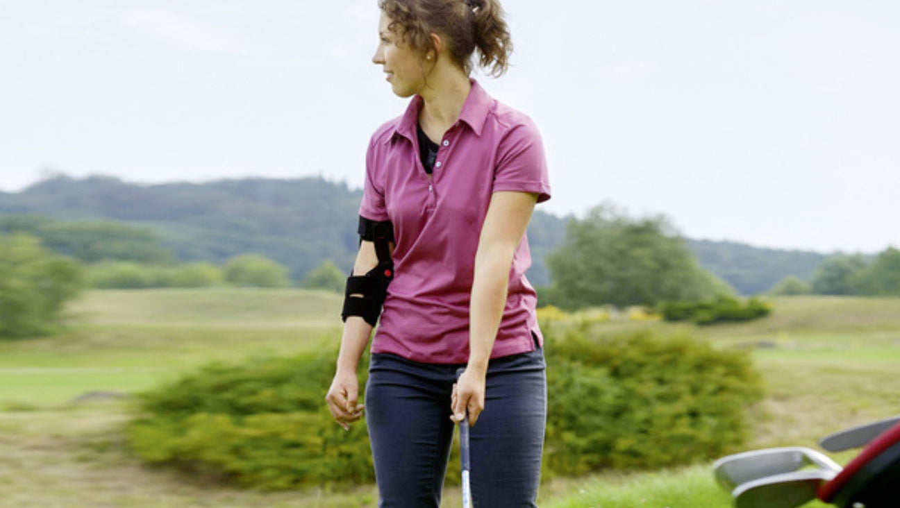 A woman wearing a pink polo shirt and arm brace stands outdoors on a golf course, holding a golf club and looking to the side. Trees and hills are visible in the background.
