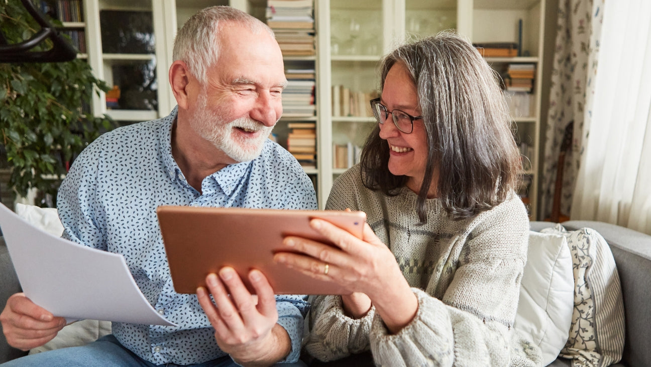 A smiling elderly couple sits on a couch, the man holding papers and the woman holding a tablet. They look at each other warmly in a cozy living room with bookshelves in the background.
