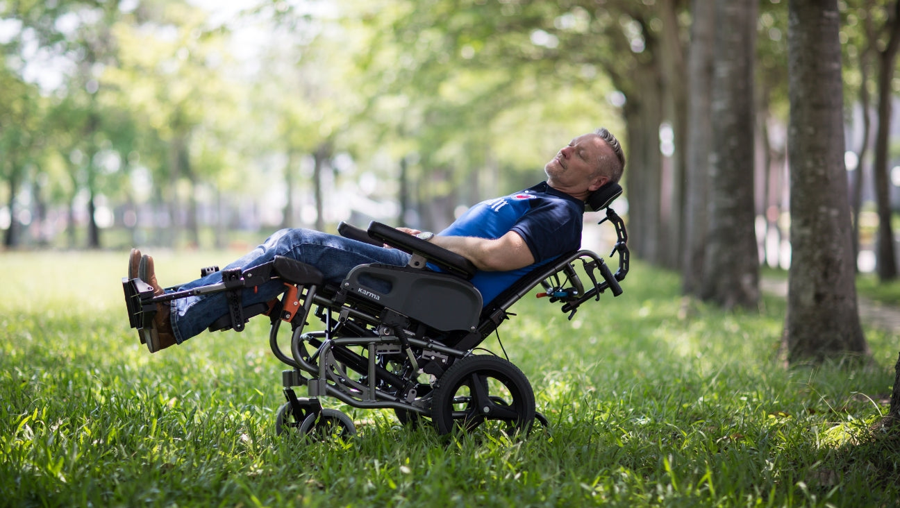 A man in a reclining wheelchair relaxes on a grassy area under trees, eyes closed, enjoying the outdoors on a sunny day.