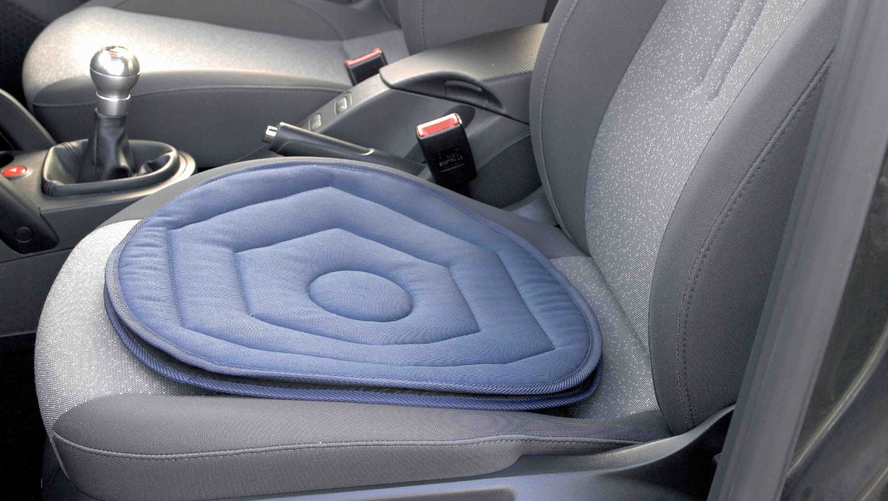 A blue hexagonal padded seat cushion is placed on the drivers seat of a car with gray fabric upholstery. The cars gear shift and seat belt buckles are also visible.