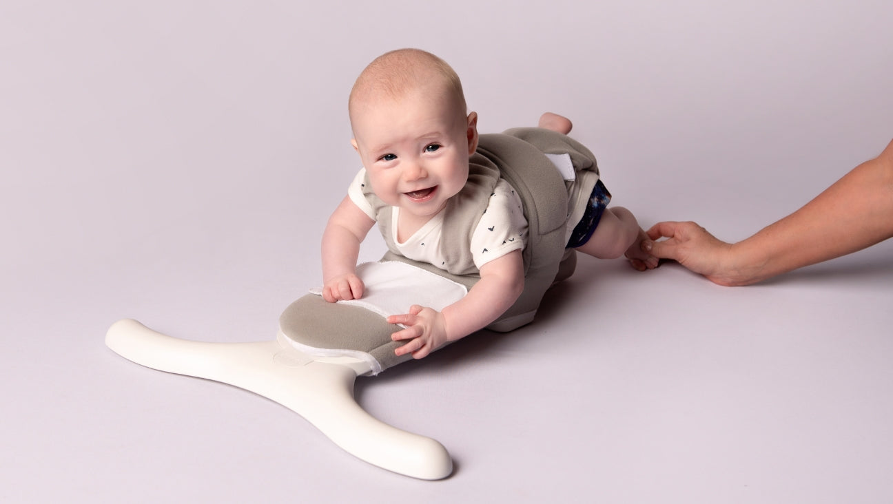 A smiling baby wearing a soft, supportive harness lies on their stomach, propped up on a white, Y-shaped device, while an adult hand gently holds the harness from behind.