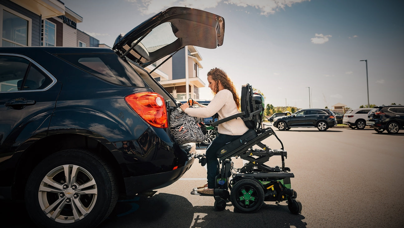 A woman in a motorized wheelchair loads a bag into the open trunk of a black SUV in a sunny parking lot, with buildings and other parked cars in the background.