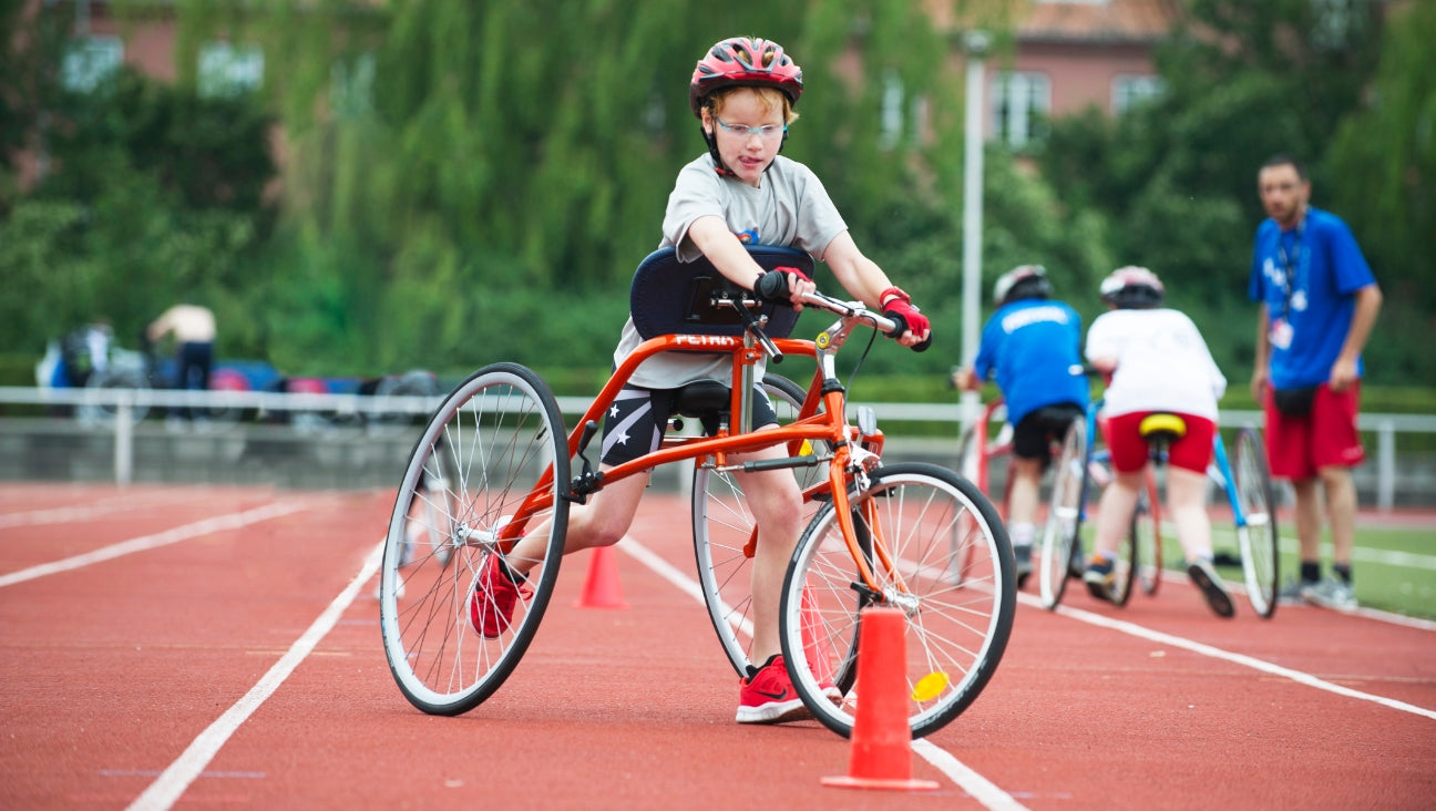 A young athlete using an adaptive tricycle maneuvers around an orange cone on a running track, wearing a helmet and sports gear during an outdoor event.