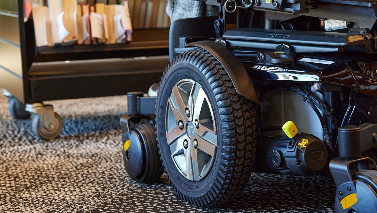 A close-up of an electric wheelchair’s wheel on a patterned carpet, with a cart filled with files in the background. The image highlights accessibility and mobility indoors.
