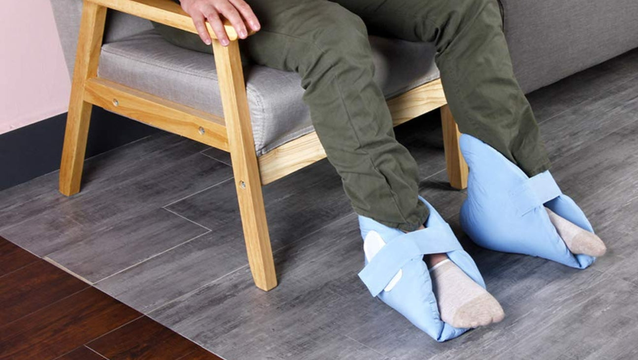 A person sitting on a wooden chair with their feet resting on the floor, wearing soft blue foot cushions secured with straps over their socks.