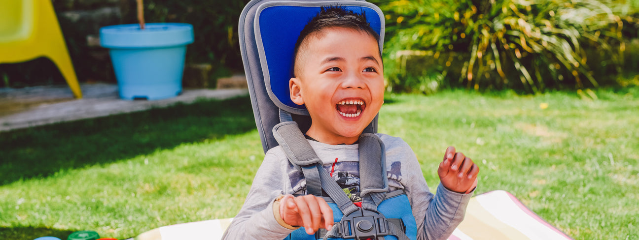 A smiling child sitting in a blue adaptive chair outdoors on a sunny day, with grass and some blurred outdoor furniture in the background.