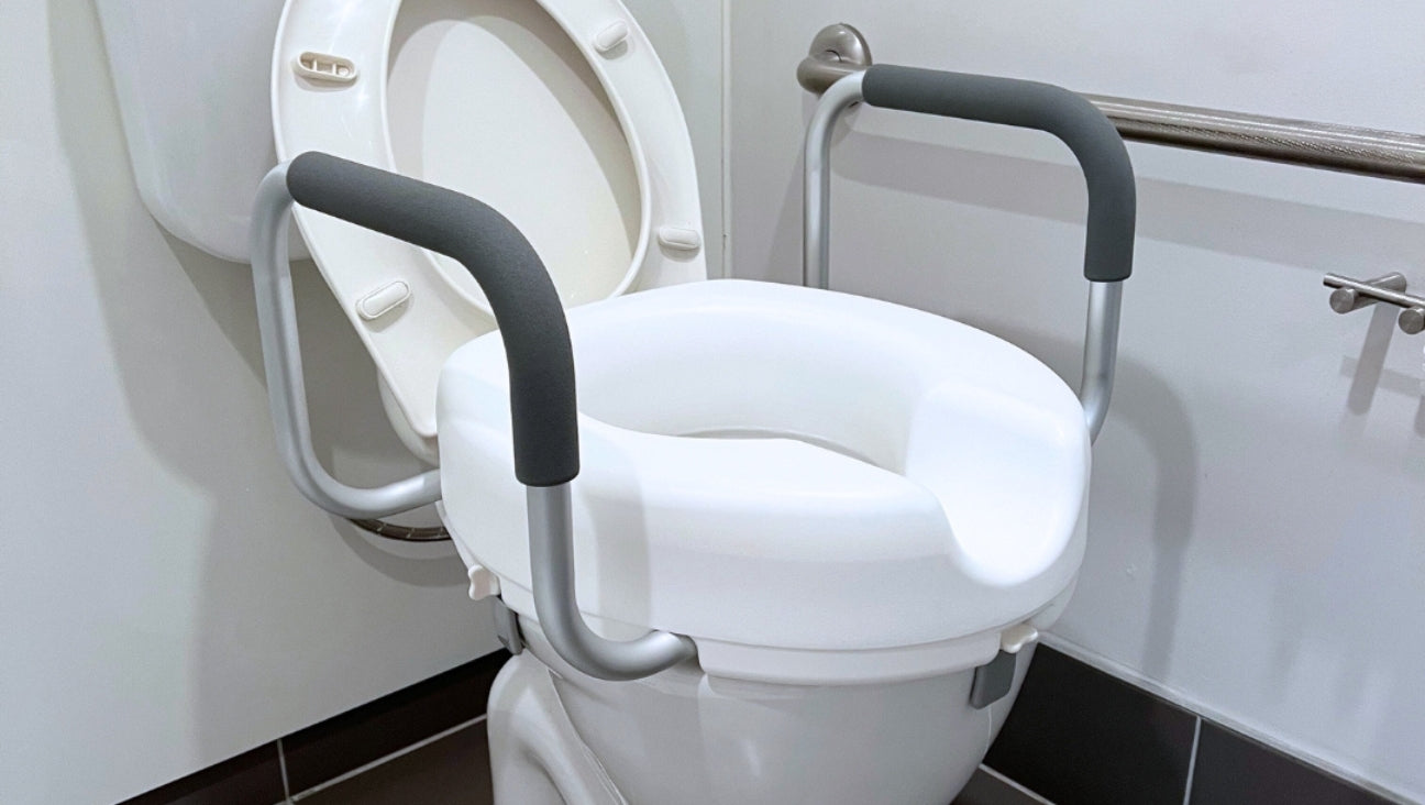 A white toilet with a raised seat attachment featuring padded armrests and a safety rail on the wall. The lid is open, and the bathroom has white walls and dark floor tiles.