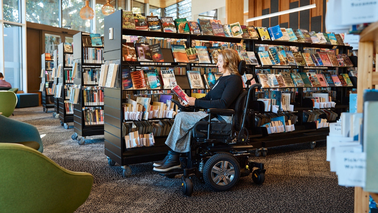 A woman in a power wheelchair reads a book in a library, surrounded by shelves filled with books and magazines. Natural light streams in through large windows in the background.