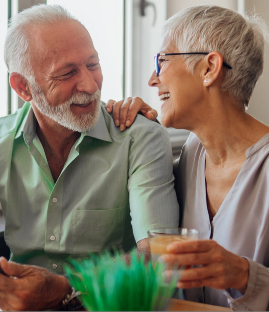 An older couple smiling and laughing together indoors. The man wears a light green button-up shirt, and the woman wears a light gray top and glasses.