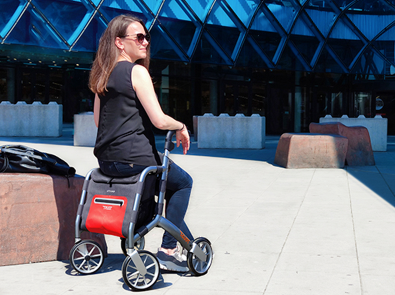 A woman in sunglasses sits on a modern red and gray rollator outdoors, with a glass building and large stone blocks in the background.
