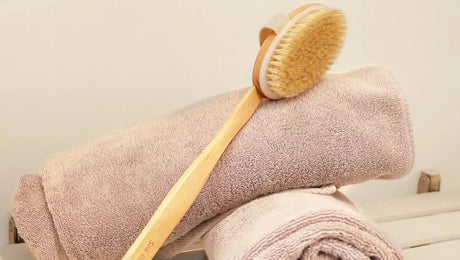 A wooden body brush rests on two neatly rolled beige towels. The soft bristles of the brush are prominently displayed. The setting suggests a spa or bathroom environment.