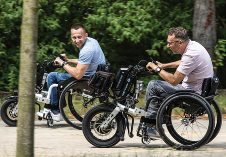 Two men in wheelchairs with front-mounted hand-bike attachments ride outdoors on a sunny day, smiling and enjoying the activity, with trees in the background.