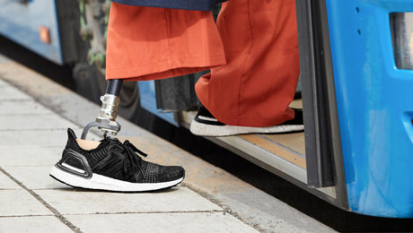 A person with a prosthetic leg, wearing orange pants and black sneakers, is stepping onto a blue bus from a sidewalk.