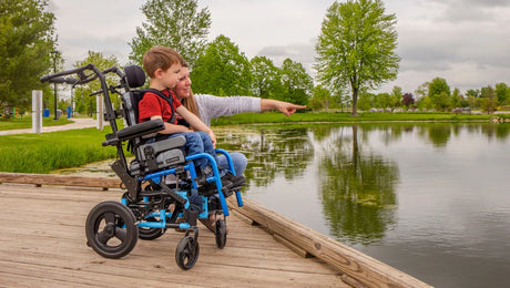 A woman and a young boy in a wheelchair sit on a wooden dock by a pond. The woman is smiling and pointing toward the water, and trees and greenery surround the calm scene.