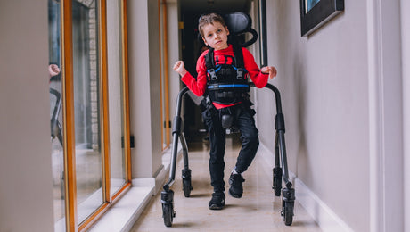A young boy using a gait trainer walks down a hallway, wearing a red long-sleeve shirt and black pants, with large windows on one side and a framed picture on the wall.