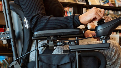 A person wearing a black sweater and gray pants uses the joystick control of a motorized wheelchair, with bookshelves visible in the background.