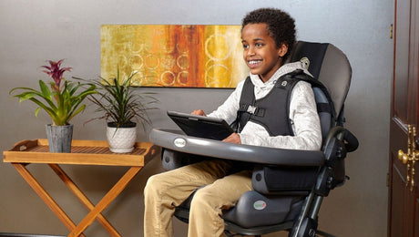 A smiling boy sits in a supportive wheelchair, using a tablet. He is indoors next to a wooden side table with potted plants and a colorful abstract painting on the wall behind him.