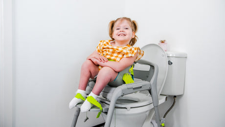 A smiling young girl with pigtails sits on a supportive chair over a toilet in a bright bathroom, wearing a yellow plaid dress, white socks, and green braces on her legs.
