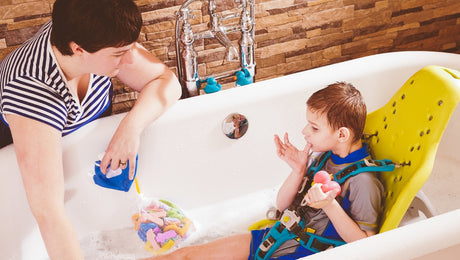 A woman helps a young boy with a disability, seated in a yellow bath chair, take a bath. The boy holds a toy while the woman uses a cup to pour water. The bathroom has brown tiled walls.