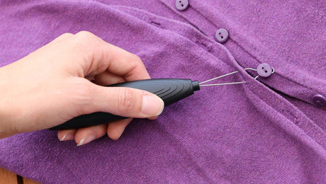 A hand uses a black sewing tool to thread a purple button onto a matching purple garment with several buttons already attached.