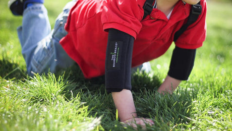 A child in a red shirt and blue jeans crawls on green grass, wearing black protective pads on their elbows. Only the lower half of the child’s face is visible.