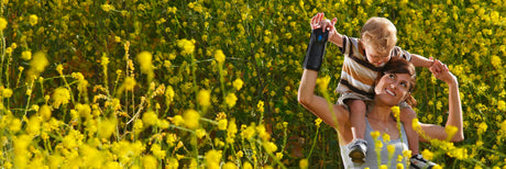 A woman carries a child on her shoulders as they walk through a field of yellow flowers. Both are smiling, enjoying the sunny day. The field stretches into the distance with vibrant blooms.