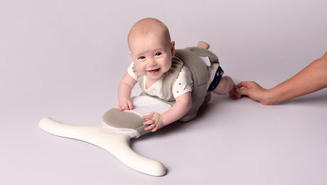 A smiling baby wearing a soft, supportive harness lies on their stomach, propped up on a white, Y-shaped device, while an adult hand gently holds the harness from behind.