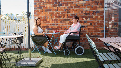 Two women sit at a small outdoor café table; one is in a wheelchair. They are smiling and chatting with drinks in front of them, next to a brick wall and surrounded by empty tables.