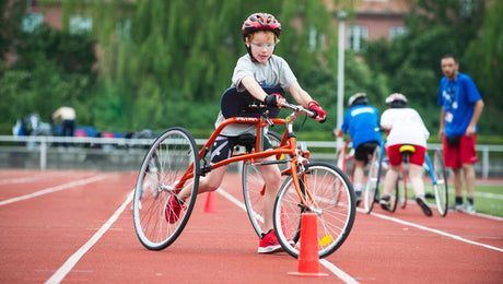 A young athlete using an adaptive tricycle maneuvers around an orange cone on a running track, wearing a helmet and sports gear during an outdoor event.