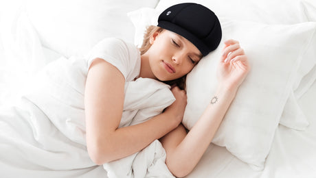 A woman sleeps on a bed with white sheets, hugging a pillow. She wears a black cap and has a small sun tattoo on her inner forearm.