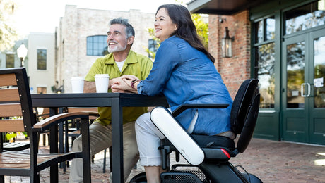 A smiling man and woman sit at an outdoor cafe table. The woman uses a powered wheelchair and both have coffee cups in front of them. They appear relaxed and are enjoying the sunny day.