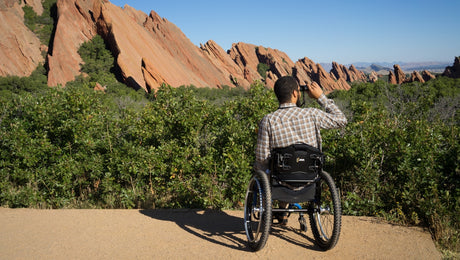 A person in a wheelchair faces red rock formations and greenery, enjoying the scenic outdoor view on a sunny day.