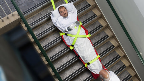 A man wrapped in a rescue sled with bright yellow straps is being transported down a staircase, assisted by another person standing at the bottom of the stairs.