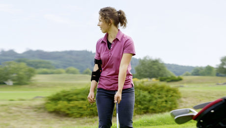 A woman wearing a pink polo shirt and an arm brace stands on a golf course, holding a golf club and looking to the side. Golf equipment and green landscape are visible.