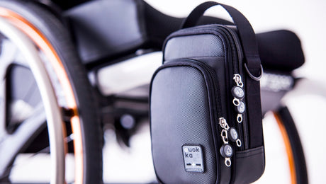 A small black bag with zippers hangs on the side of a wheelchair, with part of the wheelchair’s wheel and seat visible in the background.