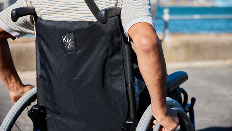 A person wearing a striped shirt uses a wheelchair outdoors near a body of water. The focus is on their hands gripping the wheels and the back of the wheelchair with the Ki logo.