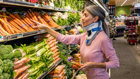 A woman wearing a neck brace shops for carrots in the produce aisle of a grocery store, reaching for a bag while holding fresh carrots in her other hand.
