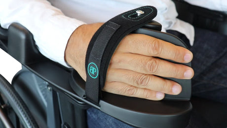 A person’s hand secured to a wheelchair armrest with a black strap, providing support and stability. The strap features a blue embroidered logo.