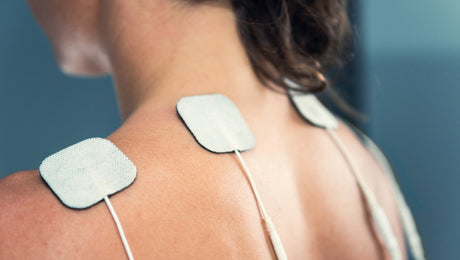 Close-up of a person’s upper back and shoulder with three adhesive electrode pads attached, connected to wires, likely for electrical therapy or pain relief treatment.