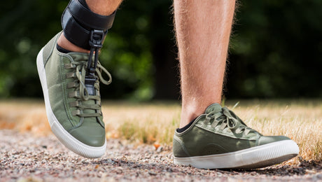 A person wearing green sneakers and an orthopedic leg brace walks on a gravel path with grass and trees in the background.