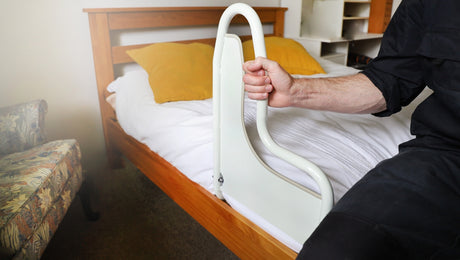 A person sits on a bed holding a white safety rail, with two yellow pillows and a floral armchair nearby.