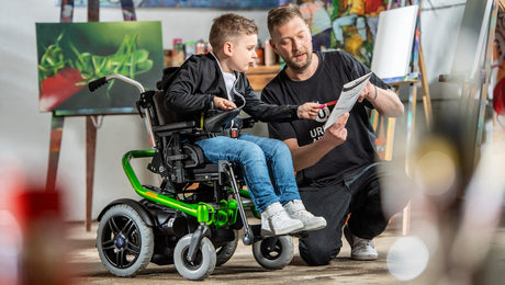 A man kneels next to a boy in a wheelchair, showing him how to draw on a pad. They are in an art studio with colorful artwork and supplies in the background.