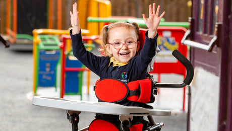 A young girl with red glasses and pigtails smiles and raises her arms while sitting in a supportive wheelchair at a colorful playground.