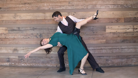 A man in formal attire dips a woman in a green dress during a dance, both smiling, against a rustic wooden wall background.
