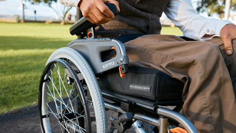 A person in brown pants sits in a wheelchair on a sunny day in a park, with one hand resting on the armrest and green grass in the background.