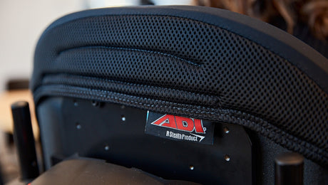 Close-up view of the backrest of a black ADI-brand chair or wheelchair, featuring a mesh-like fabric and a label reading “ADI A Stealth Product.”.