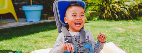 A smiling child sitting in a blue adaptive chair outdoors on a sunny day, with grass and some blurred outdoor furniture in the background.