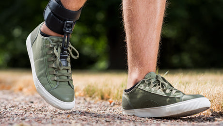 A person walks outdoors on a gravel path wearing green sneakers and an ankle-foot orthosis on one leg, with grass and trees in the blurred background.